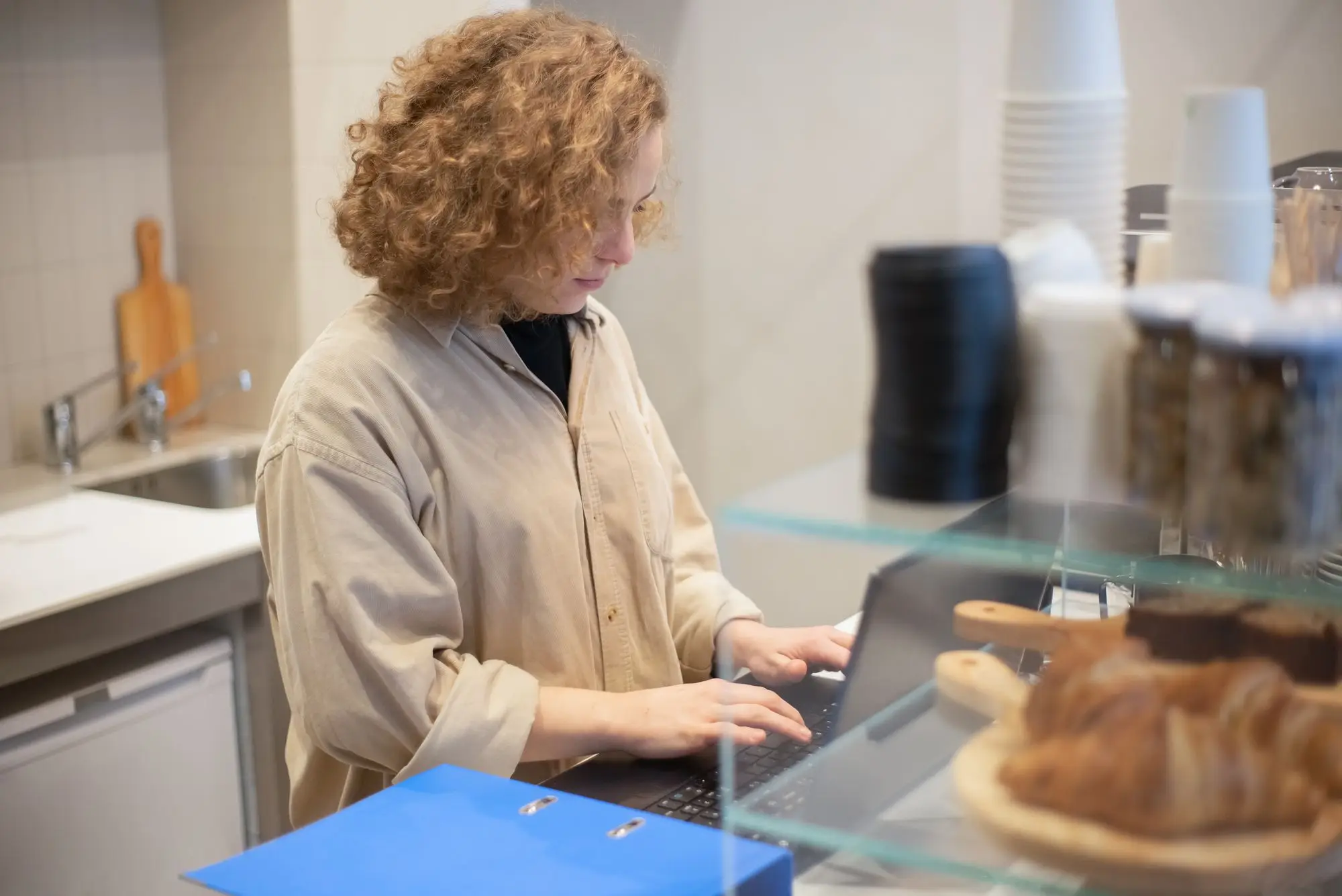 Café owner behind the counter checking an equipment rental agreement on a laptop, with a blue contract folder beside the till