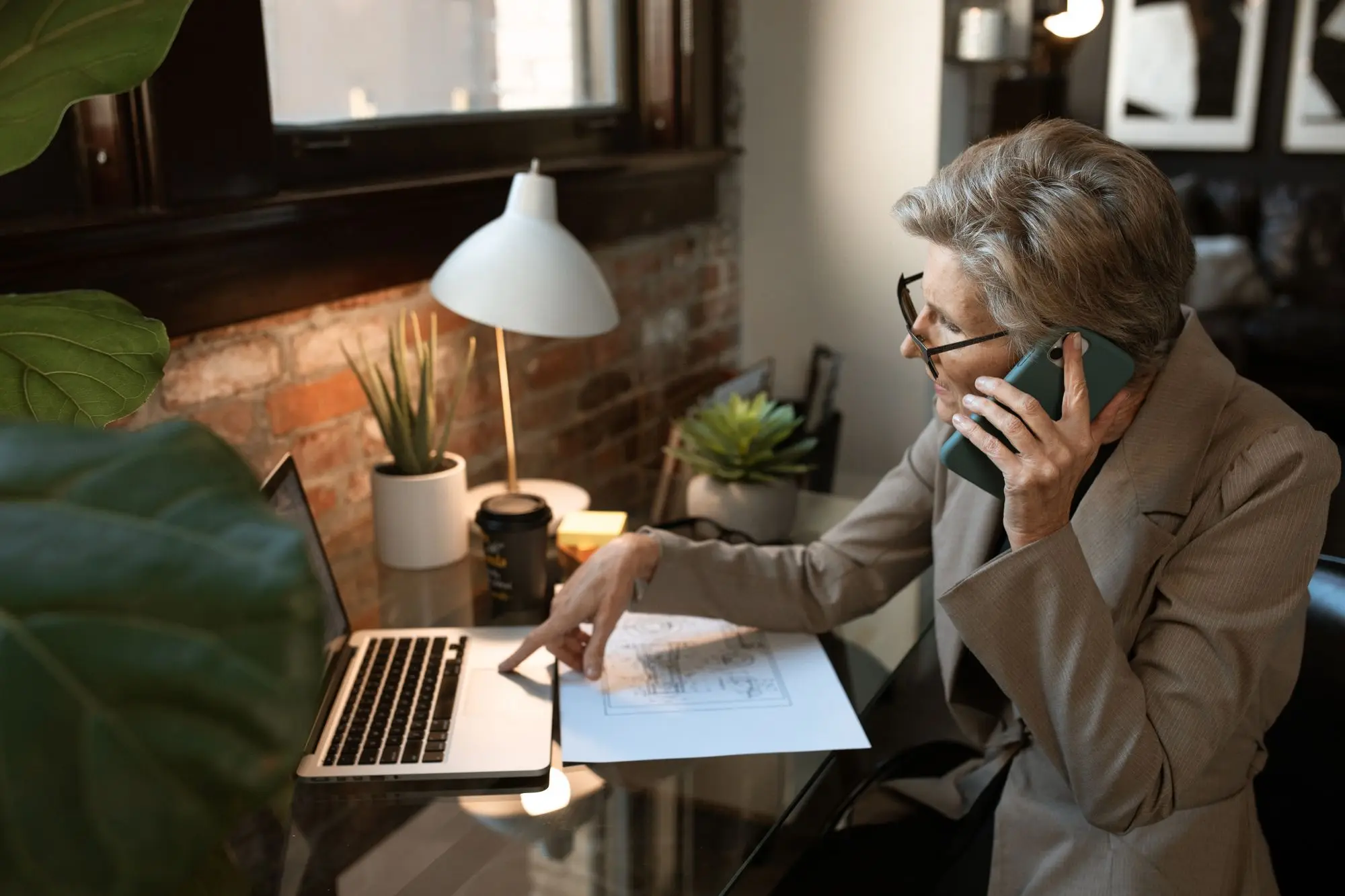 Business owner on a phone call with a lender at her desk, one hand on the printed loan offer beside her laptop